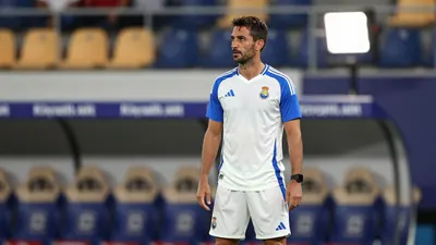 A 35-year-old Málaga CF coach, wearing a white jersey with blue sleeves and a white kit, stands in the middle of the...