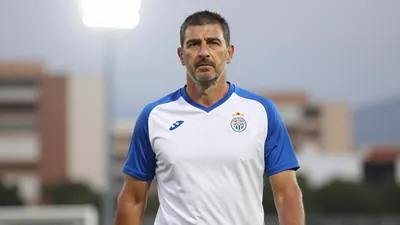 A 35-year-old male coach with a strong build, wearing a white Málaga CF jersey with blue sleeves and a blue collar...