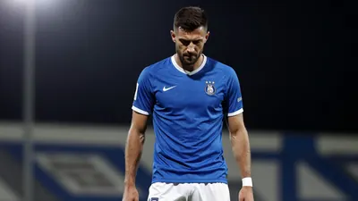 A 35-year-old Málaga CF central defender, wearing a blue jersey and white shorts, with a serious expression, looking down