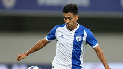 A mid-30s male midfielder in a solid blue and white kit, waist-up, focused expression, tight action crop of him striking...