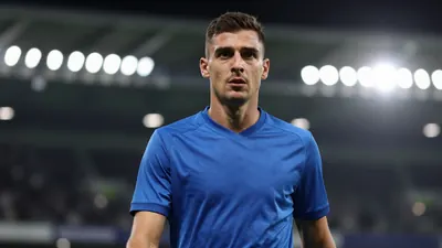 A mid-30s male central defender in a solid blue kit, waist-up, focused expression, background blurred stadium lights...