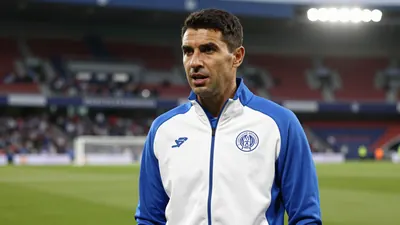 Photorealistic 85mm f/1.4 shot of a mid-30s male football manager in a white and blue tracksuit, waist-up, speaking...