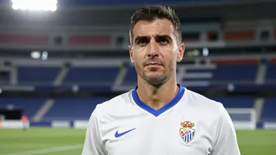 A 35-year-old male coach with a kind face, wearing a white Málaga CF jersey with a blue collar and sleeves, standing in...