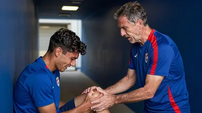A 50-year-old male athletic trainer, dressed in a plain blue and red polyester training top, stands with his back against...