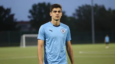 A 28-year-old centre-back with athletic build, wearing a solid light blue jersey, standing on a training pitch at dusk