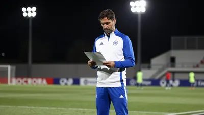 A 35-year-old Málaga CF assistant coach, wearing a white and blue tracksuit, stands in the middle of the training pitch...