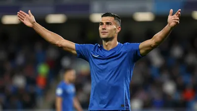 A mid-30s male midfielder in a solid blue kit, waist-up, focused expression, arms raised in celebration after scoring...