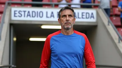 A 50-year-old, 5'10" male coach, dressed in a plain blue and red cotton training top, stands at the Estadio La Rosaleda's...