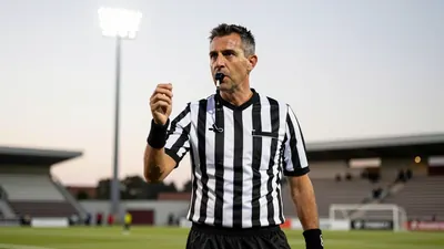 A mid-30s referee in a plain black and white striped shirt, crisp and unwrinkled, stands pitchside at Estadio La...