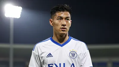 A 32-year-old midfielder in a white and blue football kit, standing on a pitch with a determined expression
