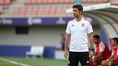 A 30-year-old Málaga CF coach, wearing a white shirt and black shorts, stands in the dugout, looking focused, with a...
