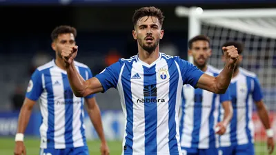 A 25-year-old Málaga CF midfielder in a blue and white striped jersey, waist-up, celebrating with teammates after scoring...