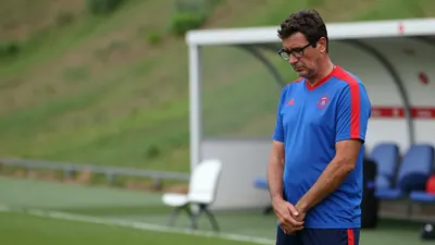 A tired, bespectacled, 30-year-old coach with a weathered, worn face, dressed in Málaga CF's blue and red plain training...
