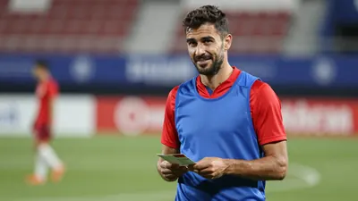 Mid-30s male Málaga CF assistant coach, athletic build, waist-up, wearing an unmarked training top split blue left side...