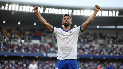 Photorealistic image of a Málaga CF player in a white jersey and blue shorts, celebrating with arms raised in La Rosaleda...
