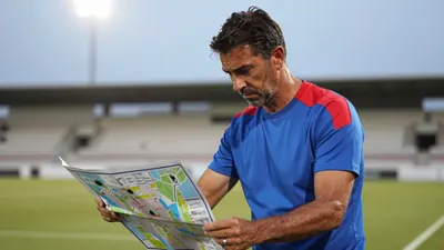 A medium shot of a rugged, 35-year-old, athletic coach, dressed in a plain, blue and red, cotton training top, with a...