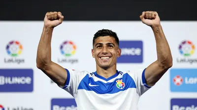 A Málaga CF player in a white and blue jersey with a broad smile, celebrating with arms raised, standing on a stage...